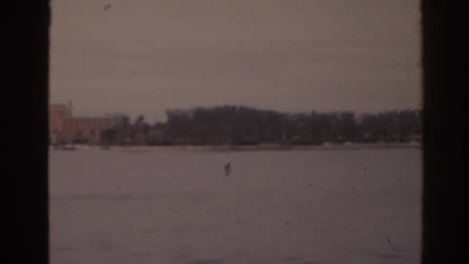 1963 Florida home-movie frame showing calm water, bare trees, and a low building on the far shore.