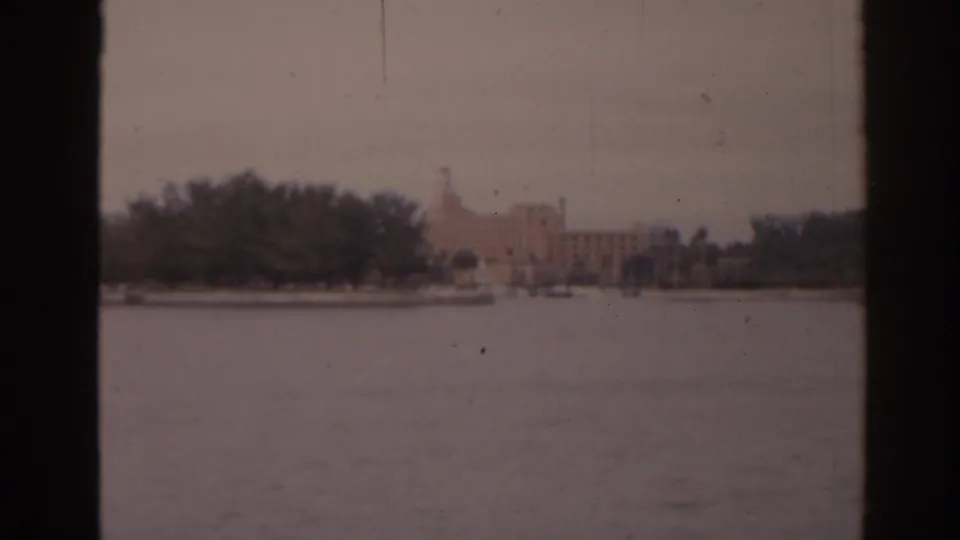 1963 Florida 8mm view across water toward a pale shoreline building with a tower, trees, and dark boat-window edges.