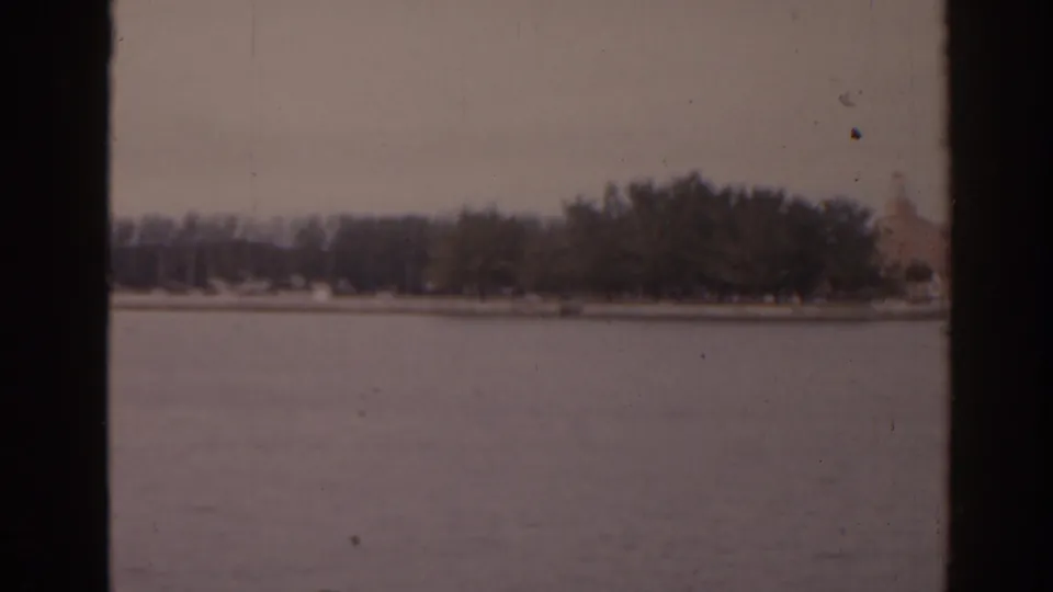 1963 Florida home-movie frame showing water, a tree-covered shore, and part of a pale building near the edge of view.