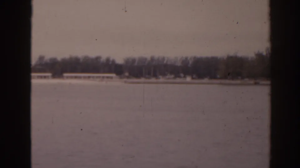 Soft 8mm view across a Florida waterway toward trees and a long pale shoreline structure under a gray sky.