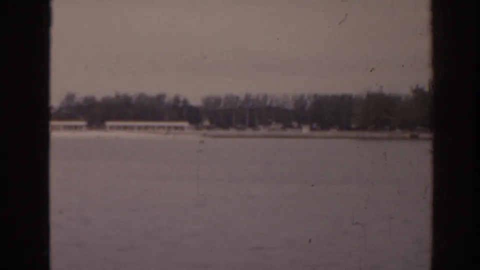 1963 Florida 8mm frame showing a distant tree-lined shore and long low buildings across a pale lake or waterfront surface.