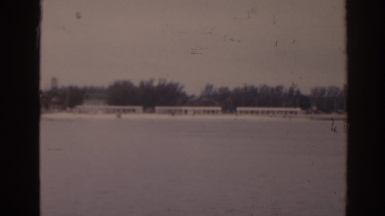 Distant 1963 Florida waterfront view with a tree line, low shore buildings, and heavy dark frame edges from the 8mm scan.