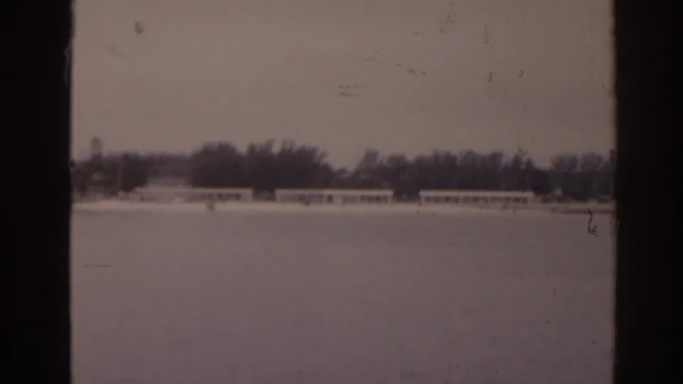 Distant 1963 Florida waterfront view with a tree line, low shore buildings, and heavy dark frame edges from the 8mm scan.