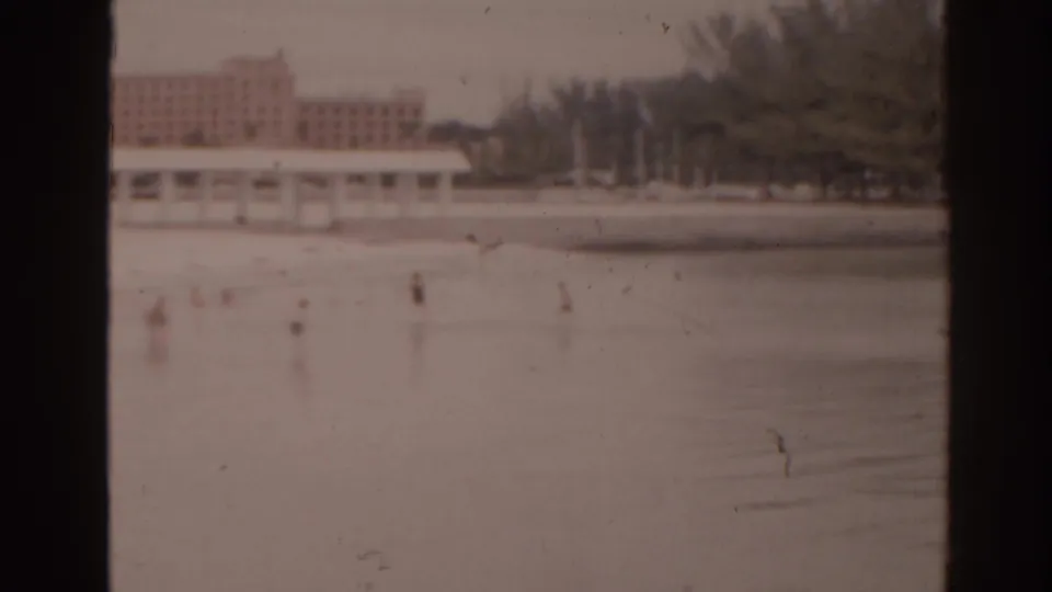1963 Florida home-movie frame with small distant people on a pale waterside surface, long low buildings, trees, and a larger building behind them.