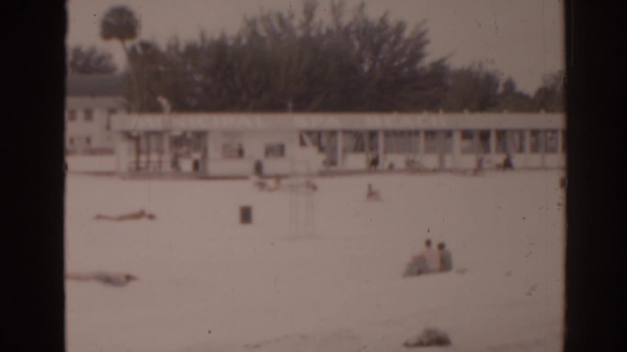 Distant beachgoers on pale sand near a low building with dark film borders.