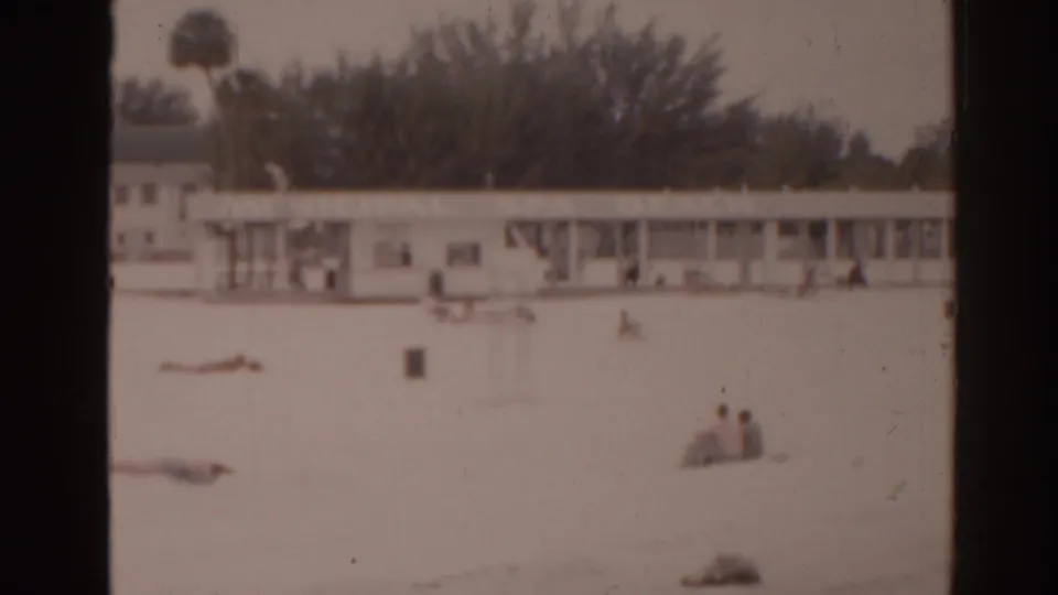 1963 Florida 8mm frame showing a quiet beach with distant people resting on sand and a low beachfront building beyond.