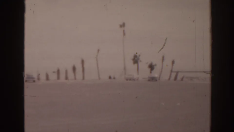 Soft 8mm Florida beach frame with parked cars, tall palms, and small distant figures on open sand.