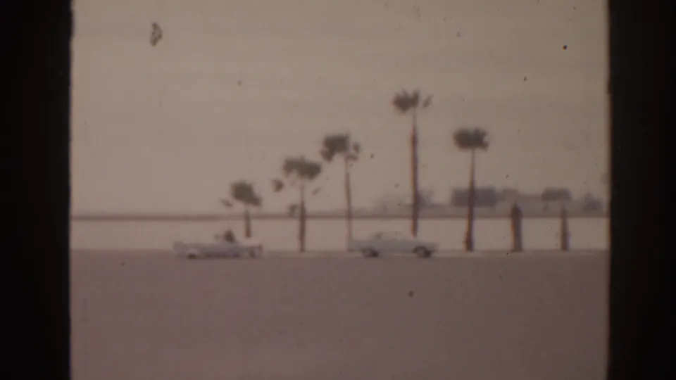 1963 Florida 8mm frame showing cars under palm trees with pale beach, bright water, and a hazy horizon.