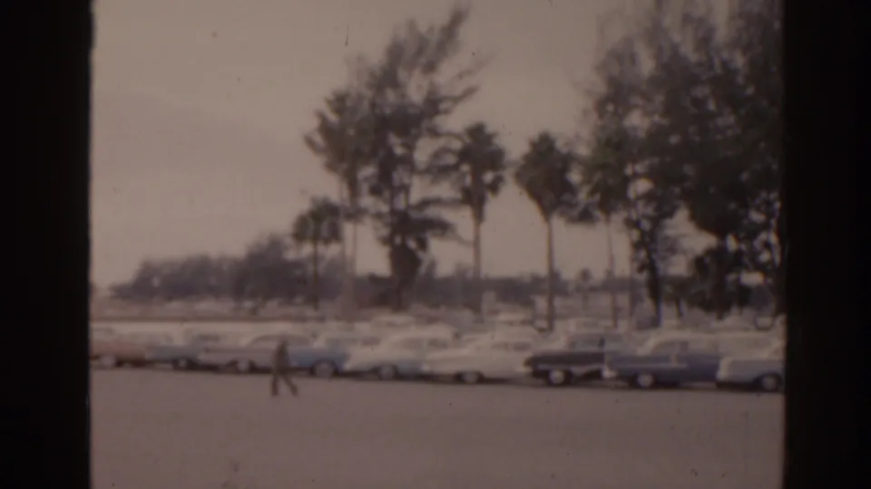 1963 Florida 8mm frame with a lone distant figure crossing the lot, parked cars, palms, and dense greenery behind.