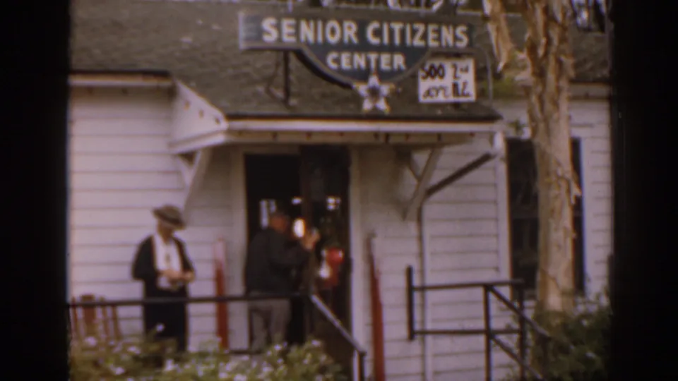 1963 Florida home-movie frame showing two adults near the entrance of a white clapboard Senior Citizens Center with readable signage.
