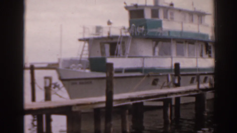 1963 Florida waterfront frame showing a large moored boat beside a wooden dock, with pilings and water in the foreground.