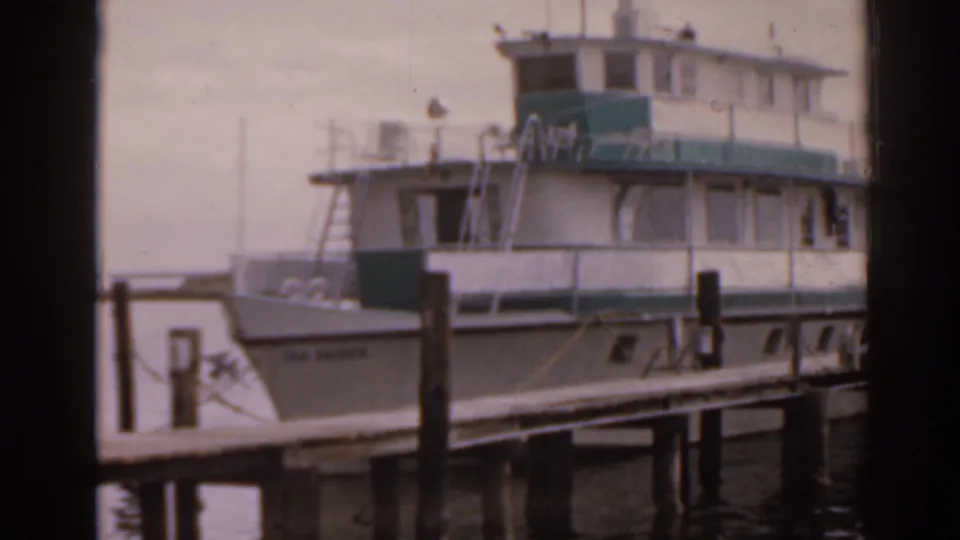 1963 Florida home-movie frame of a moored waterfront boat near dock pilings, with soft focus and dark frame edge visible.