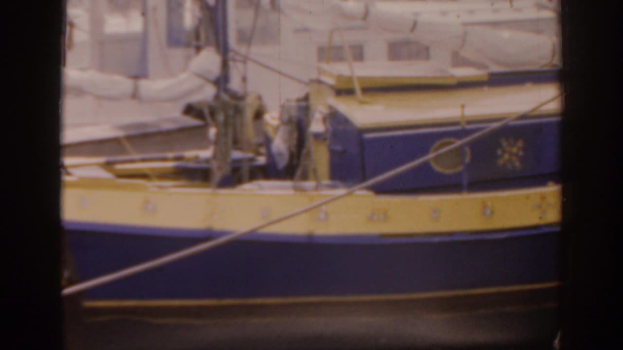 The camera frames the boat's blue hull and yellow band from the pier, with dock lines, hardware, and cabin windows in soft 8mm focus.
