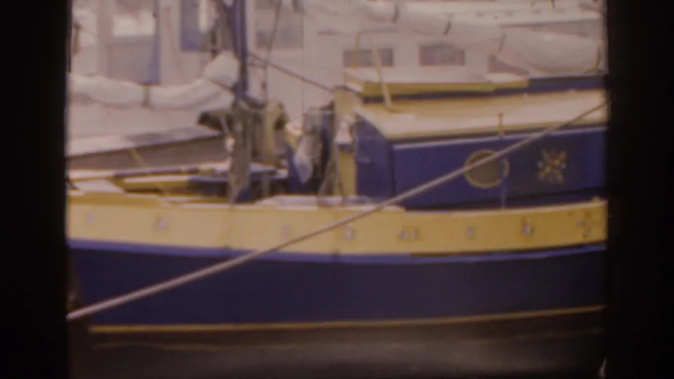 The camera frames the boat's blue hull and yellow band from the pier, with dock lines, hardware, and cabin windows in soft 8mm focus.