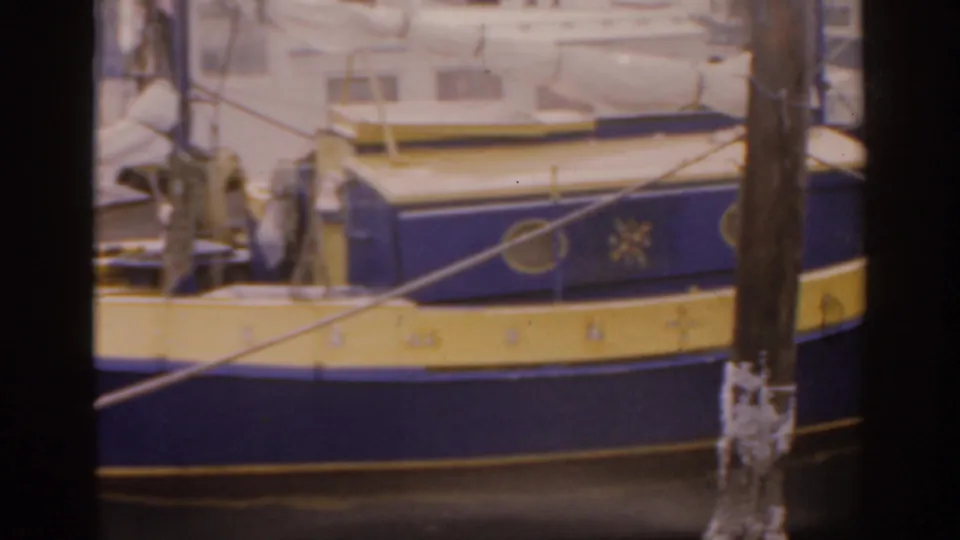 A close 1963 Florida home-movie view shows the boat's painted hull behind a weathered pier post, with rope and water reflections below.