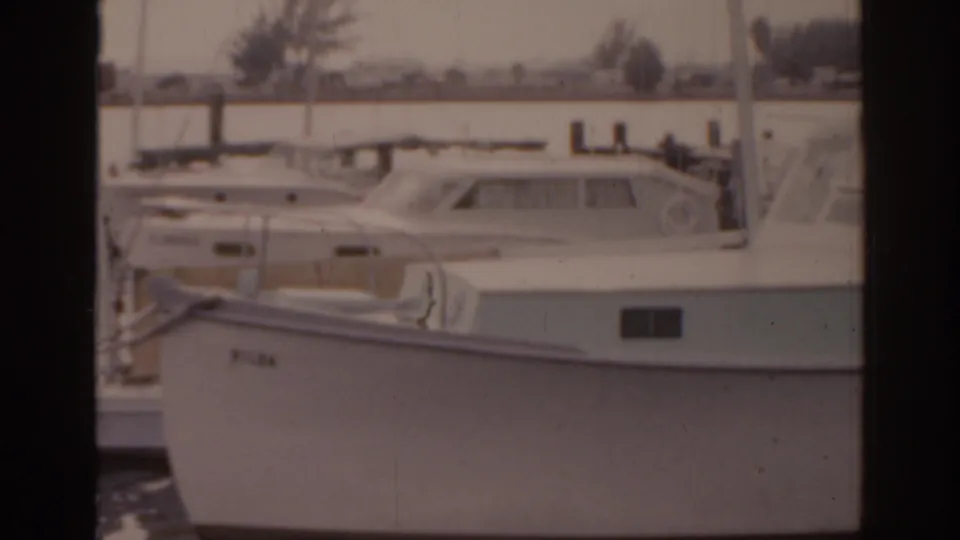 1963 Florida 8mm frame showing a larger white cabin boat alongside a dock, with calm water and shoreline in the background.