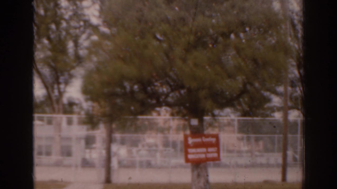 1963 Florida 8mm frame showing trees, a chain-link fence, and a red Tampa Adult Education Center sign across a quiet street.