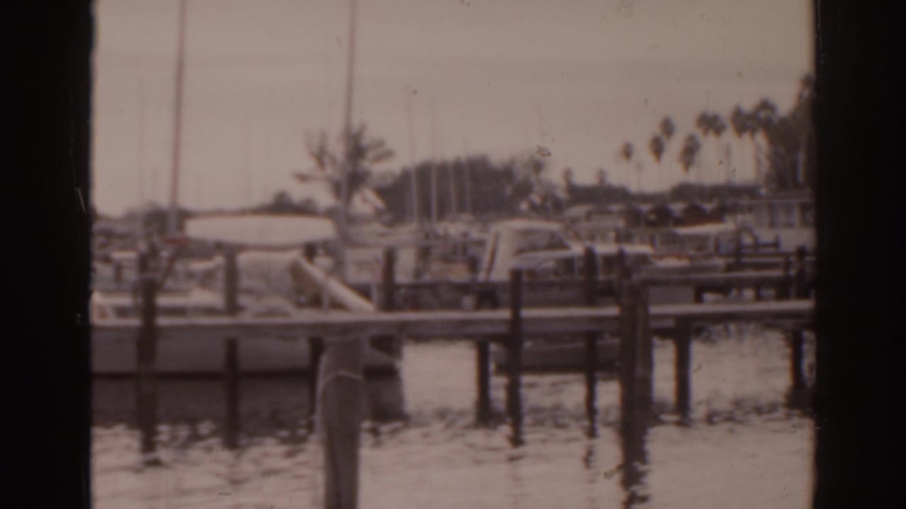 1963 Florida 8mm frame of boats tied along wooden marina piers, with calm water, palms, and small human figures in the background.