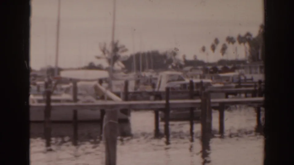 1963 Florida 8mm frame of boats tied along wooden marina piers, with calm water, palms, and small human figures in the background.