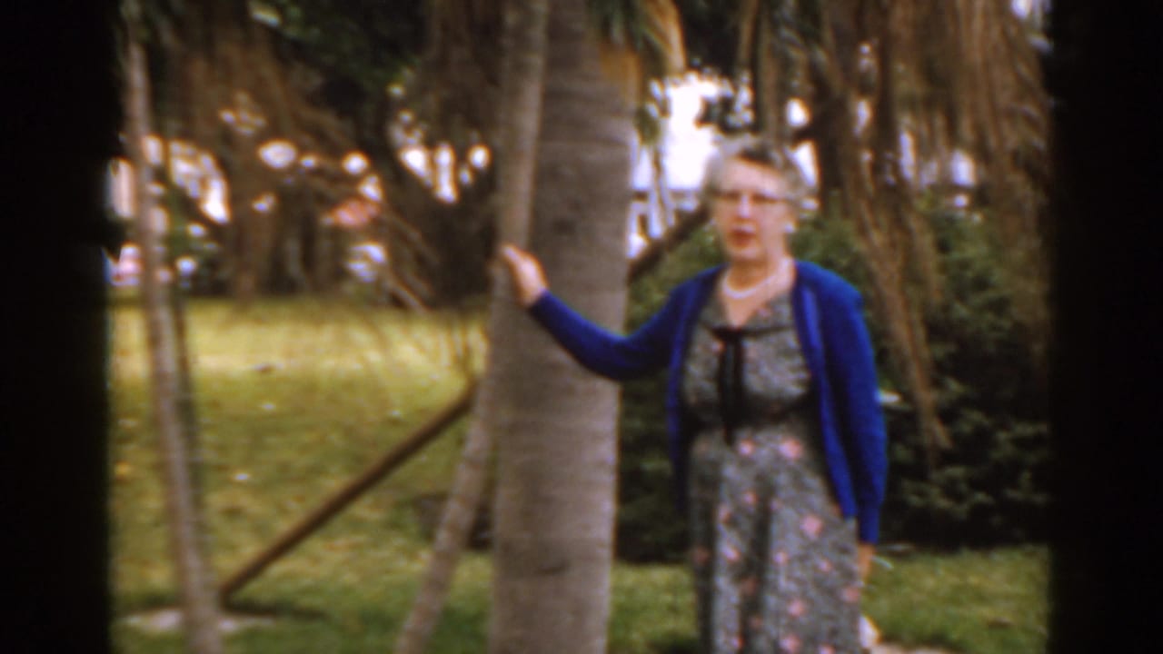Mature woman in a blue sweater stands beside a tree in a green park.
