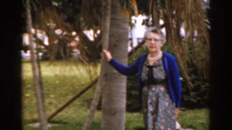 The 1963 Florida home-movie frame shows a mature woman in glasses, floral dress and blue cardigan leaning against a tree.