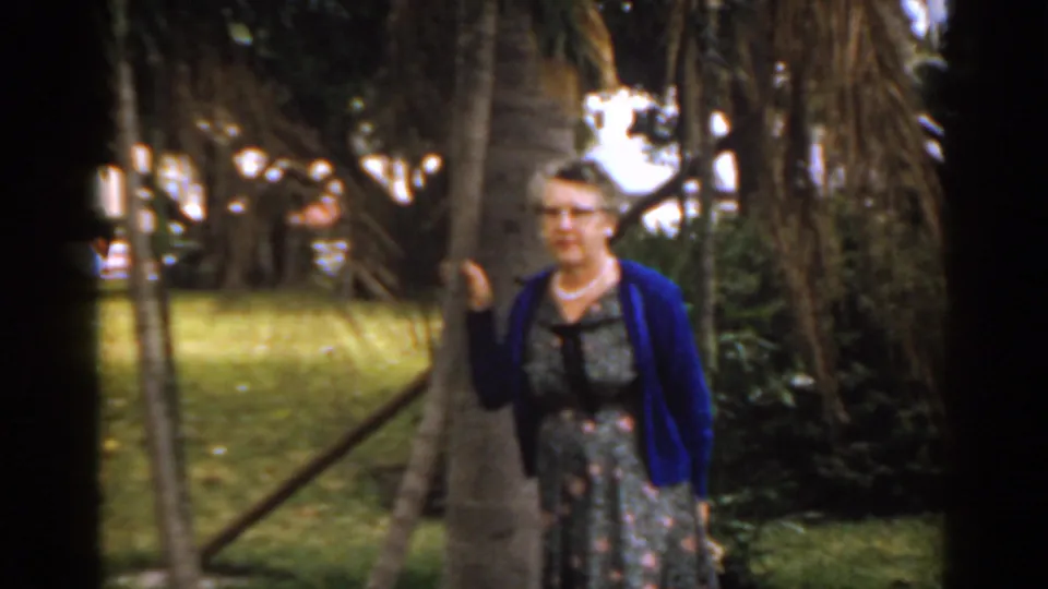 A mature woman in a blue cardigan and floral dress stands beside a tree in a leafy Florida park, facing the camera.