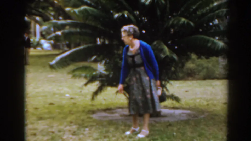 Older woman in 1963 Florida garden footage looks toward a low plant, framed by broad tropical leaves and shaded grass.