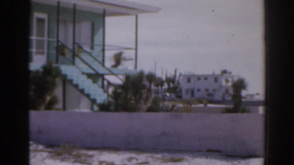 1963 Florida beachfront frame showing the stair side of the turquoise building with palms, a low wall, and a pale building beyond.