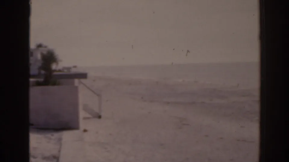 1963 Florida 8mm view of a wide sandy beach, low wall, sparse palm silhouette, and hazy ocean horizon.