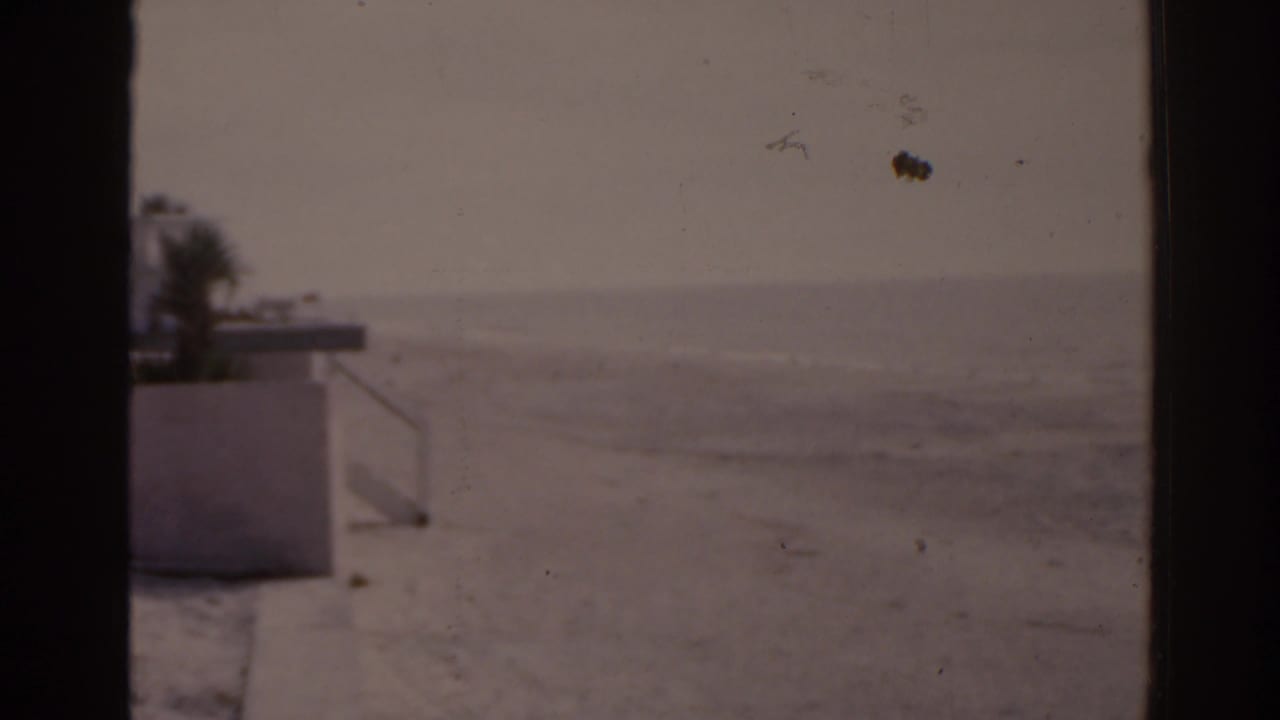 Empty sandy beach with ocean horizon, framed by dark film borders and a concrete structure at left.