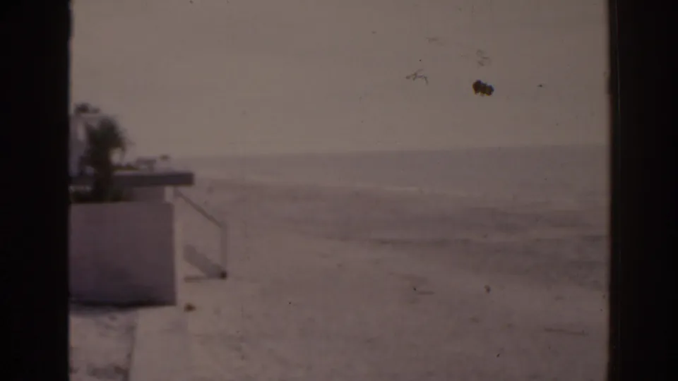 1963 Florida 8mm view of an empty beach and distant ocean, with a low concrete structure and palm foliage at the left edge.