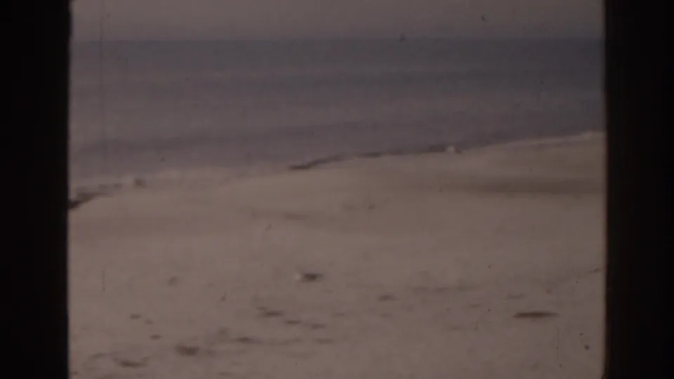 1963 Florida shoreline with gentle waves and open sand, partially obscured by dark 8mm frame edges.
