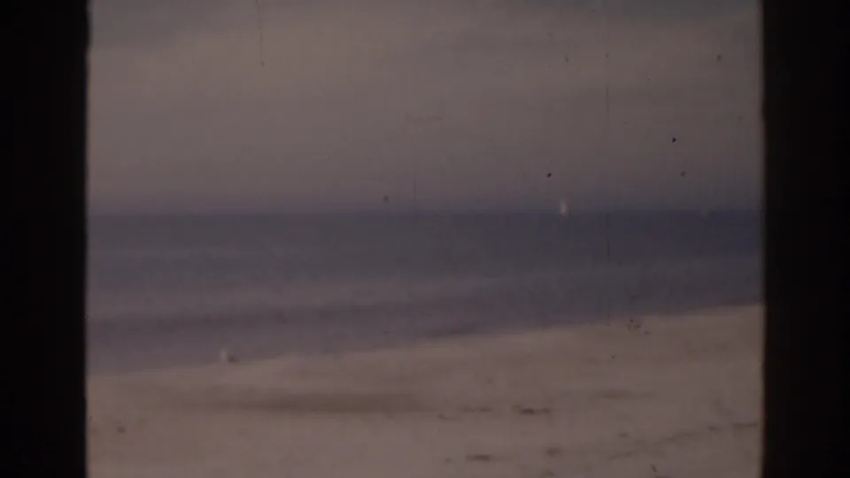 1963 Florida beach view with pale sand, gentle surf, muted sky, and visible 8mm frame edges.