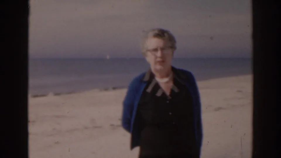 Elderly woman with glasses stands on a Florida beach near the water, filmed in soft 8mm color.