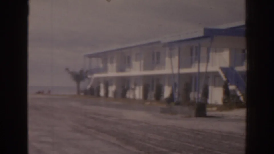 1963 Florida 8mm view of a two-story roadside motel-like building with a blue awning, exterior stairs, and an empty road.