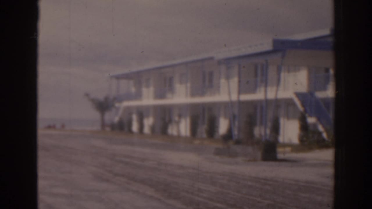1963 Florida 8mm frame showing a low white roadside building, exterior stair, pavement, and a white parked car under muted cloudy light.
