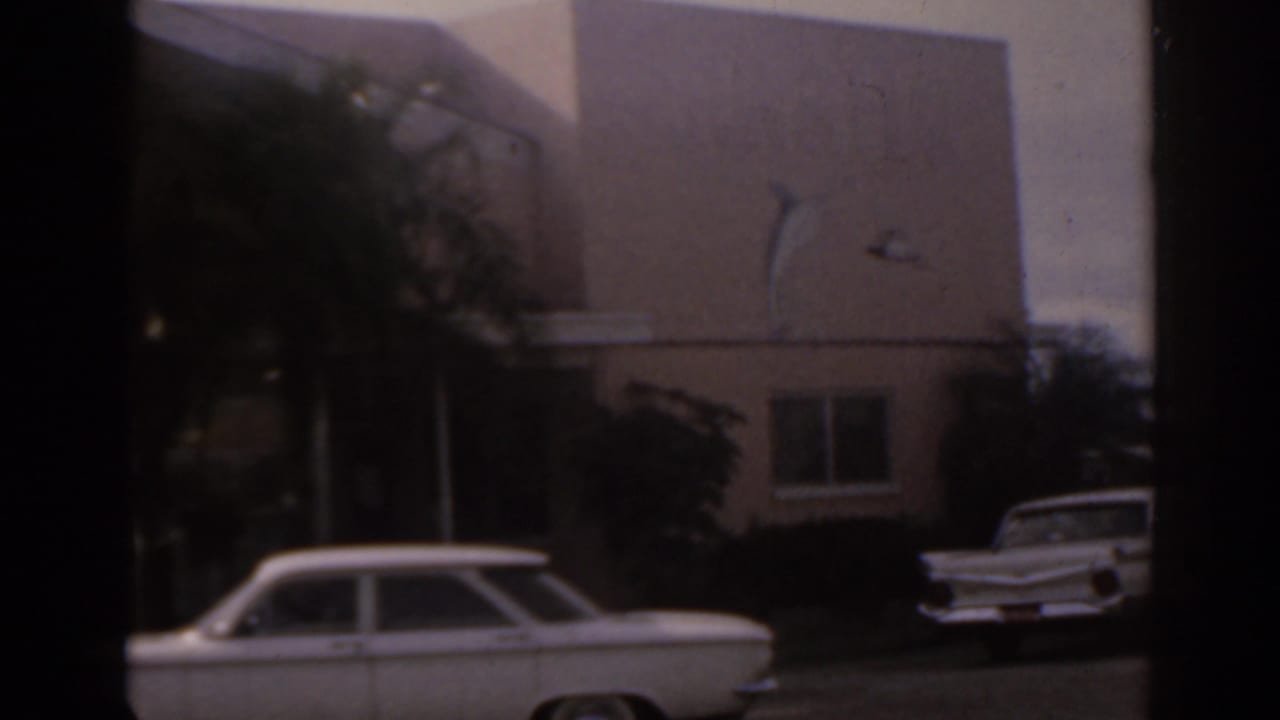 1963 Florida home-movie frame with a white station wagon outside a tan building, palm plantings, and a second white car partly visible.