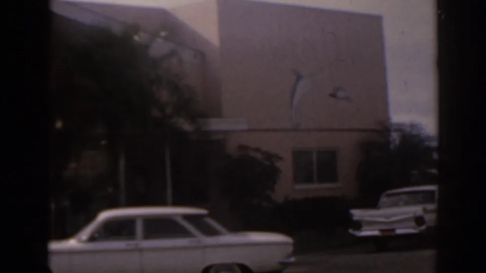 1963 Florida home-movie frame with a white station wagon outside a tan building, palm plantings, and a second white car partly visible.