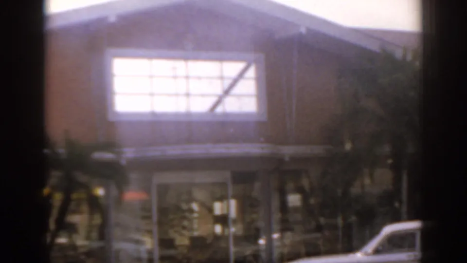 1963 Florida 8mm frame showing a tan building with a large bright upper window, palm trees, glass doors, and part of a white car.