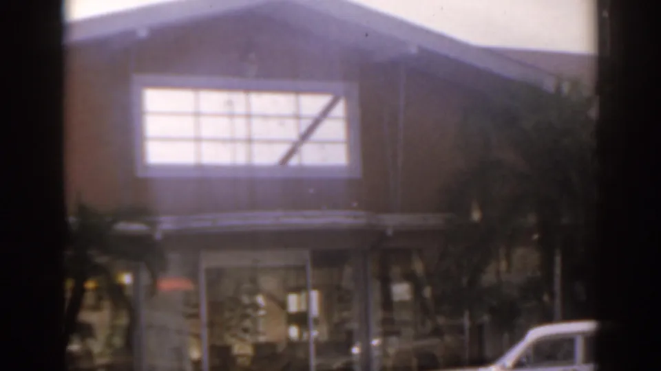1963 Florida home-movie frame repeating the tan building exterior with a large window, palms, glass entry, and a cropped white car at lower right.