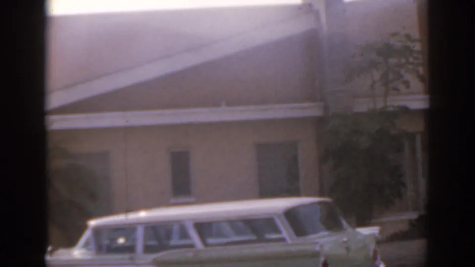 1963 Florida home-movie frame with a white station wagon beside a low building and a sunlit avenue beyond.
