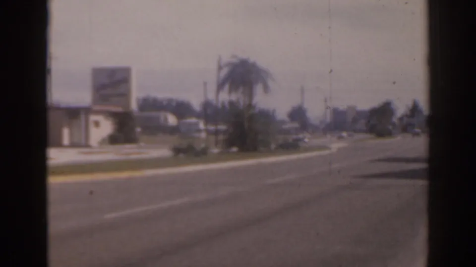 Faded 8mm frame of a 1963 Florida avenue with palm trees, low roadside buildings, and faint distant traffic.