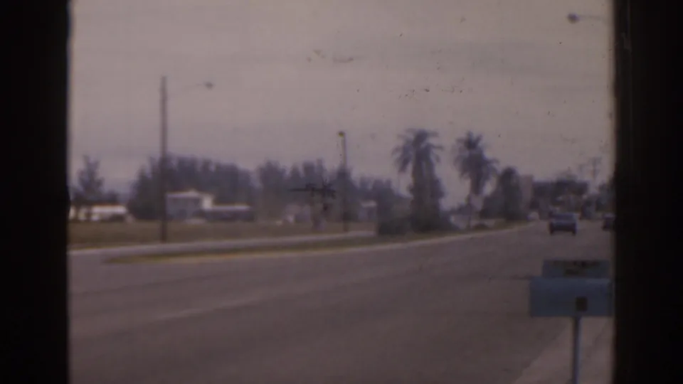 1963 Florida home-movie frame showing a broad roadside view with palm trees, utility poles, distant houses, and light traffic.