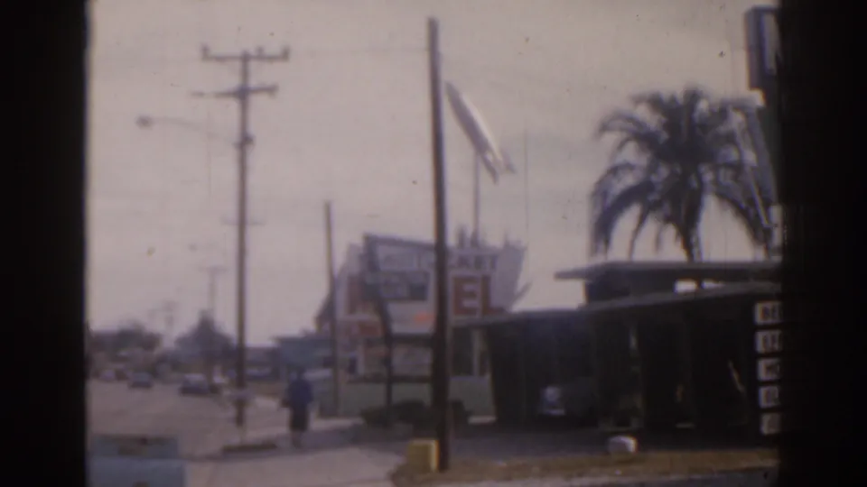 1963 Florida roadside frame with a motel-style sign, palm tree, flag, utility poles, passing cars, and a distant pedestrian.