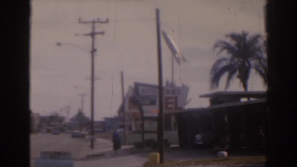 1963 Florida home-movie frame showing a motel-style sign, palm tree, flag, utility poles, roadway traffic, and a distant person on the sidewalk.