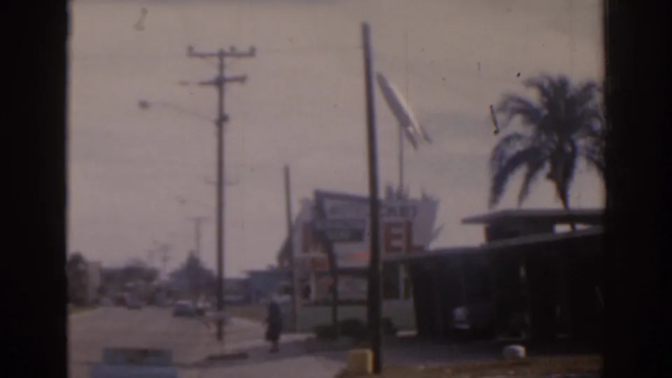 1963 Florida roadside scene with utility poles, motel signage, palm trees, parked cars, and a person walking near the sidewalk.