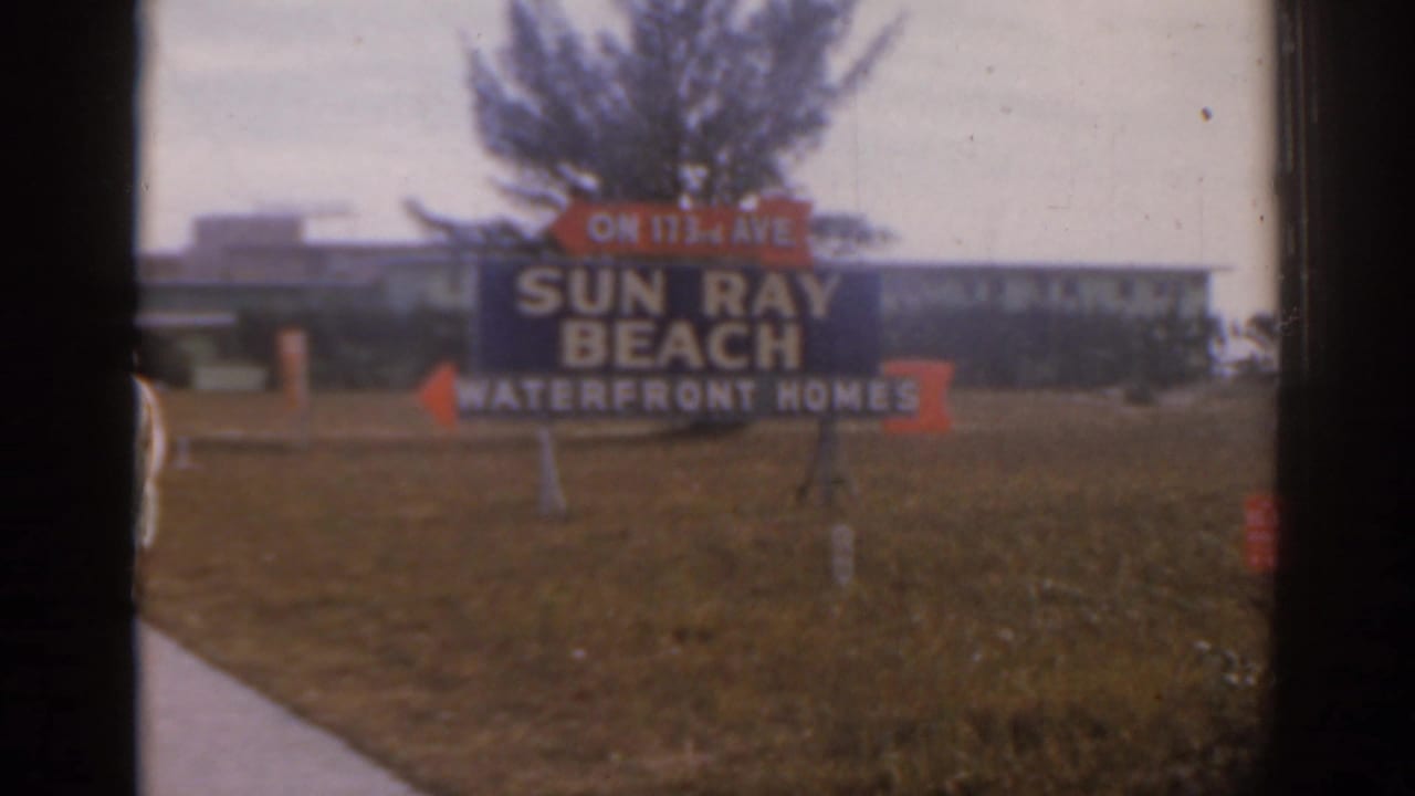 Sun Ray Beach Waterfront Homes sign beside a sidewalk, lawn, palm tree, and low building.