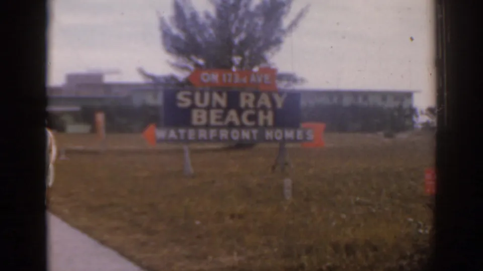 Florida roadside sign reading Sun Ray Beach Waterfront Homes with a sidewalk, lawn, palm tree, and low-rise building behind it.