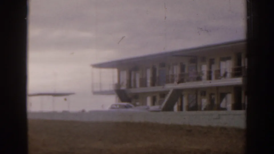 1963 Florida view of a low-rise waterfront motel or apartment building with balconies, exterior stairs, parked cars, lawn, and water in the distance.
