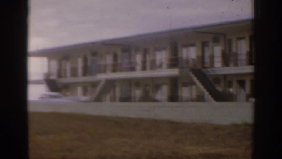 Florida coastal building with exterior staircases, balcony walkway, parked cars, grass foreground, and a partial view toward the water.